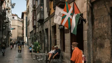 Calle estrecha con gente, mesas al aire libre y banderas ondeando en un ambiente urbano.