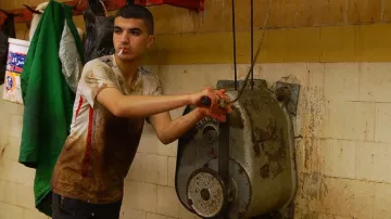 L'image montre un jeune homme dans un environnement de travail, probablement une boucherie ou un lieu de préparation alimentaire. Il est habillé de manière assez sales, avec des taches sur son t-shirt. Il tient un outil ou une manette pour actionner une machine, et il a une cigarette à la bouche. L'arrière-plan est constitué de murs carrelés avec des traces de saleté, créant une ambiance industrielle. L'atmosphère semble intense et le travail semble exigeant.