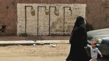 L'image montre une femme vêtue d'une tenue noire, probablement un hijab ou une burqa, marchant dans une rue. À ses côtés, un enfant tient sa main. En arrière-plan, on aperçoit un mur de béton avec un graffiti qui représente des pots de peinture suspendus, ajoutant une touche artistique à la scène. La route semble modeste avec des débris au sol, ce qui donne un aperçu de l'environnement urbain.