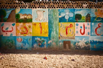 The image shows a wall decorated with colorful murals or paintings. The artwork includes various designs such as flowers, shapes, and possibly symbols or letters that are arranged in a grid pattern. The colors are vibrant, featuring shades of blue, green, yellow, and pink, giving the wall a lively and artistic feel. The surrounding area appears dusty or sandy, suggesting an outdoor environment. Overall, the scene conveys a sense of creativity and community.