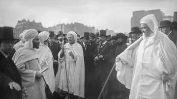 L'image montre un groupe de personnes dans un environnement urbain. Au centre, plusieurs individus portent des vêtements traditionnels, tels que des robes blanches et des turbans. D'autres, habillés de manière formelle avec des chapeaux haut de forme et des costumes, se tiennent autour d'eux, suggérant une rencontre entre différentes cultures. L'arrière-plan présente des bâtiments typiques de l'époque. L'atmosphère semble être celle d'un événement officiel ou d'une cérémonie.