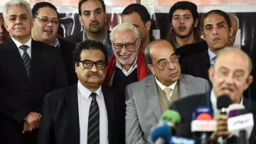 The image features a group of people gathered together, possibly at an event or press conference. There are several men in formal attire, with some wearing suits and ties, while others seem to be casually dressed. In the foreground, one man is speaking into a microphone, while others appear to be listening attentively. There is a mix of expressions among the individuals, including smiles and serious looks. The background includes a banner, suggesting this may be a significant gathering related to a specific topic or announcement.