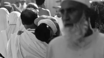 L'image montre une scène animée d'une foule, probablement lors d'un événement religieux. On peut voir un homme portant un enfant sur son dos, tandis que d'autres personnes sont visibles en arrière-plan, certaines portant des vêtements traditionnels. L'image est en noir et blanc, ce qui accentue l'atmosphère solennelle de l'événement. Les expressions des visages et les attitudes des personnes semblent refléter une ambiance de dévotion et de spiritualité.