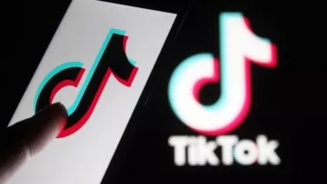 The image features a close-up of a smartphone displaying the TikTok logo, which is designed with vibrant colors—predominantly red, blue, and white. A finger is pointing towards the logo on the screen, while a larger version of the TikTok logo is blurred in the background. The overall focus is on the application's recognizable branding.