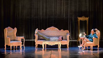 L'image montre une scène de théâtre avec un décor composé de plusieurs meubles anciens. Au centre, un sofa sur lequel un homme est allongé, semble détendu. À droite, une femme est assise sur une chaise, elle semble engagée dans une conversation ou une réflexion. Le fond est constitué d'un rideau sombre, ce qui met en valeur les meubles éclairés. L'ambiance semble à la fois intime et dramatique, suggérant une interaction entre les deux personnages.