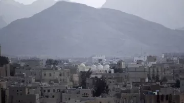 L'image montre un paysage urbain avec une vue sur des bâtiments dans une ville entourée de montagnes. On peut voir quelques structures avec des dômes, indiquant peut-être des mosquées ou des monuments. Le décor est dominé par des tons de gris et un ciel nuageux, créant une atmosphère sombre et mélancolique. Les montagnes au loin ajoutent une dimension naturelle à la scène, contrastant avec l'urbanisation.