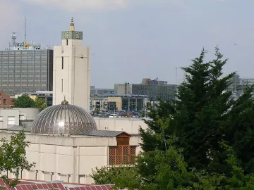 L'image montre un bâtiment architectural avec un dôme et une tour horloge, entouré d'arbres et d'autres structures urbaines en arrière-plan. On peut voir un environnement mixte, combinant des éléments d'architecture moderne et traditionnelle, avec des stations de transports et des bâtiments de bureaux. La scène est bien éclairée, suggérant une journée ensoleillée.