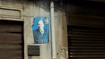 L'image montre un mur d'un bâtiment avec une affiche décolorée d'un homme qui semble être une figure publique. L'affiche est légèrement abîmée, avec des déchirures et des marques. À côté, il y a une plaque signalétique en arabe décorative, elle-même fixée au mur, qui semble indiquer le nom d'un établissement ou d'une personne liée au bâtiment. Le mur présente également des signes d'usure, avec une peinture écaillée et quelques éclats de surface. Les volets en métal, qui sont fermés, ajoutent à l'atmosphère urbaine de la scène.