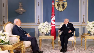 L'image montre deux hommes assis dans un cadre officiel, probablement dans un bureau ou une salle de réunion élégante. Les deux sont vêtus de costumes sombres, l'un portant des lunettes. Au fond, on peut voir le drapeau tunisien ainsi qu'un blason. La pièce est décorée avec des meubles en or, des fleurs blanches et un tapis coloré, ce qui crée une atmosphère formelle et importante.