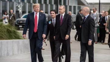 L'image montre un groupe d'hommes en costumes, apparemment dans un cadre formel, probablement lors d'un événement diplomatique. Deux hommes, respectivement en costume noir et rouge, marchent côte à côte, tandis que d'autres les suivent. L'arrière-plan indique un environnement urbain moderne, avec des véhicules et des bâtiments. L'atmosphère semble sérieuse, éventuellement liée à des discussions ou des négociations importantes.