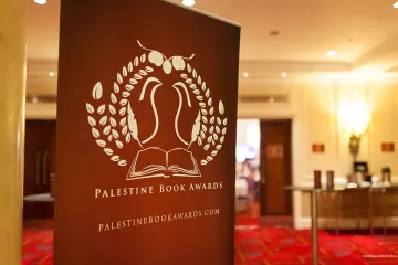 L'image représente un panneau de présentation pour les Palestine Book Awards. Ce panneau arbore un design avec des éléments graphiques, incluant une illustration d'un livre ouvert entouré de feuilles. Le texte indique "Palestine Book Awards" ainsi que le site web associé. L'arrière-plan semble être une salle de réception, avec un sol tapissé et des éclairages doux.