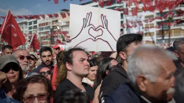 L'image montre une grande foule rassemblée lors d'un événement en plein air. Au centre, une personne tient une pancarte avec un dessin de deux mains formant un cœur. La majorité des personnes présentes portent des vêtements aux couleurs nationales, notamment du rouge et du blanc, et certains affichent des drapeaux turcs. L'ambiance semble festive et engagée, témoignant d'une manifestation ou d'une célébration collective.