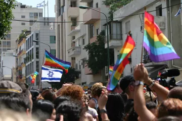 L'image montre une scène vibrante de célébration lors d'un défilé de fierté. On y voit une foule de personnes brandissant des drapeaux arc-en-ciel, symboles de la communauté LGBTQ+. Parmi eux, une grande bannière représentant le drapeau d'Israël flotte également. En arrière-plan, on distingue des bâtiments urbains, et plusieurs personnes semblent s'amuser et célébrer l'événement, créant une ambiance festive et joyeuse.