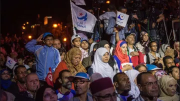 L'image montre une foule rassemblée lors d'un événement, probablement politique. On peut voir des personnes de différents âges, y compris des enfants. Certaines personnes portent des drapeaux avec des symboles, suggérant qu'elles soutiennent un mouvement ou un parti. L'atmosphère semble intense, avec des caméras et des journalistes présents, indiquant l'importance de l'événement. Les gens affichent diverses expressions, allant de la concentration à l'enthousiasme.