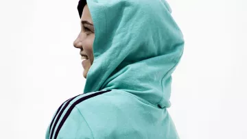 L'image montre une personne de profil, portant un sweat à capuche de couleur turquoise. Le sweat arbore le mot "QATAR" inscrit à l'arrière. La personne semble sourire, et la capuche est relevée sur sa tête. L'arrière-plan est neutre, mettant l'accent sur la tenue de la personne.