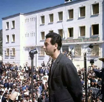 L'image montre un homme se tenant derrière un microphone, semblant s'adresser à une grande foule rassemblée devant lui. L'arrière-plan présente un bâtiment de style architectural simple avec plusieurs fenêtres, possiblement dans un contexte historique ou politique. Les membres de la foule semblent attentifs à son discours. L'éclairage naturel donne une ambiance claire à la scène, suggérant qu'elle pourrait avoir été prise en plein jour.