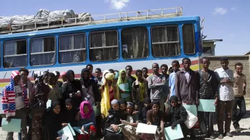 L'image montre un groupe de jeunes adultes et d'adolescents rassemblés devant un bus coloré. Ils semblent être en train de poser pour la photo, tenant tous des documents ou des fichiers. Les personnes portent des vêtements variés, incluant des hijabs et des tenues traditionnelles. L'environnement semble être en plein air, avec un ciel clair et quelques structures visibles en arrière-plan. L'atmosphère paraît conviviale et joyeuse.