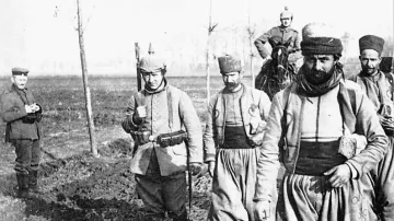 L'image montre un groupe de soldats marchant sur un terrain boueux. Ils portent des uniformes traditionnels, comprenant des pantalons larges et des bonnets. L'un des soldats est monté sur un cheval, tandis qu'un autre se tient en retrait, à l'écart du groupe. Le décor semble rural, avec quelques arbres en arrière-plan, suggérant un contexte militaire historique. L'atmosphère est sérieuse et solennelle, reflétant les conditions de la guerre.