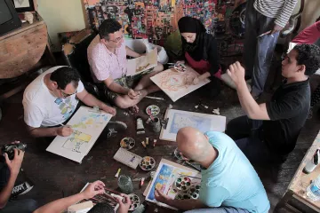 L'image montre un groupe de personnes assises sur le sol dans un espace créatif. Elles semblent engagées dans une activité artistique, utilisant des pinceaux et de la peinture sur des feuilles de papier. Certains participants semblent discuter ou partager des idées, tandis que d'autres se concentrent sur leur travail. L'environnement est coloré, avec des œuvres d'art visibles sur les murs et une palette de peinture au centre. L'ambiance paraît conviviale et collaborative.