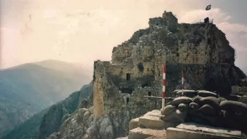 L'image montre les ruines d'une vieille forteresse perchée sur une colline, avec des murs en pierre partiellement érodés. Des drapeaux flottent au sommet de la structure. En premier plan, on peut voir des sacs de terre disposés en protection, suggérant une utilisation militaire. La vaste vallée et les montagnes se dessinent à l'arrière-plan, créant une ambiance de paysages montagneux majestueux. L'ensemble évoque une atmosphère historique et stratégique.