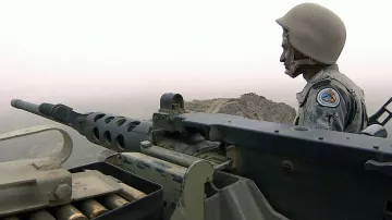 The image depicts a soldier sitting beside a mounted machine gun, which is positioned for observation or potential engagement. The soldier is wearing a helmet and a military uniform adorned with insignia. The background features a foggy or hazy landscape, suggesting a possibly mountainous or rugged terrain. The overall atmosphere conveys a sense of vigilance and readiness in a military setting.