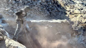 The image depicts a worker operating a pneumatic drill in a rocky area. The person is wearing protective clothing, including a mask or scarf over their face to shield against dust. Surrounding the worker are loose rocks and debris, and a cloud of dust is being kicked up by the drilling activity, creating a hazy atmosphere. The setting appears to be a mining or construction site.