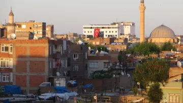 L'image montre un paysage urbain avec un mélange de bâtiments de différentes tailles et styles. On peut voir des toits plats, des antennes paraboliques et des structures en brique. À l'arrière-plan, il y a une mosquée avec un grand dôme et un minaret. La lumière du soleil crée une ambiance dorée, et quelques arbres verdoyants sont visibles, ajoutant une touche de nature à cette scène urbaine.