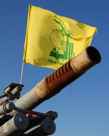 L'image montre un canon pointant vers le ciel, avec un drapeau jaune flottant au-dessus. Sur le drapeau, il y a des inscriptions en arabe ainsi qu'un logo symbolique. Le fond est bleu, suggérant une journée ensoleillée. Le canon présente des signes d'usure, avec une surface rouillée.