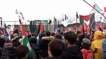 L'image montre une foule rassemblée lors d'une manifestation. Des personnes tiennent des drapeaux de différentes couleurs, dont des drapeaux palestiniens et d'autres symboles politiques. On peut voir une grande structure en arrière-plan, probablement une barrière ou une clôture, avec des bannières accrochées. L'atmosphère semble dynamique et engagée, reflétant une ambiance de protestation ou de revendication. Le temps paraît nuageux, ajoutant à l'intensité de l'événement.