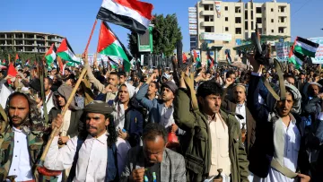 The image depicts a large crowd of people gathered in an outdoor setting, likely during a protest or rally. Many individuals are holding flags, including ones featuring a red, white, and black design, which suggests a political or national theme. The crowd appears energized, with some participants raising their fists or flags. In the background, there are buildings, indicating an urban environment. Overall, the scene conveys a strong sense of collective action and expression.