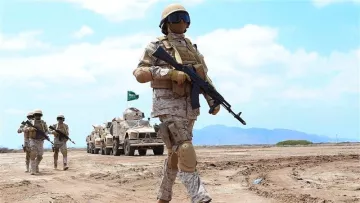 L'image montre des soldats en tenue militaire marchant sur un terrain désertique. Ils portent des casques et des gilets pare-balles, et certains utilisent des armes. En arrière-plan, on peut voir des véhicules militaires. Le ciel est clair, et le paysage est aride. La scène évoque une ambiance de formation ou d'exercice militaire.