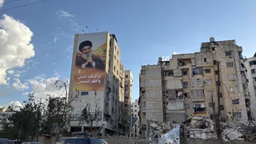 L'image montre un bâtiment en ruines, probablement à la suite d'un conflit ou d'une catastrophe, avec un autre bâtiment voisin encore debout mais en mauvais état. Sur le mur du bâtiment en meilleur état, il y a une grande affiche représentant un homme, probablement un leader ou une figure politique, tenant un symbole de pouvoir. Le ciel est partiellement nuageux, ce qui donne une ambiance contrastée à la scène. Les structures montrent des signes de délabrement, avec des fenêtres manquantes et des balcons en désordre.