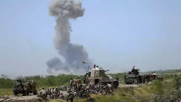 L'image montre un paysage militaire avec des soldats au sol et des véhicules blindés. En arrière-plan, une épaisse colonne de fumée s'élève dans le ciel, suggérant une explosion ou un feu. Les soldats semblent se regrouper, certains assis au sol, tandis que d'autres sont postés près des véhicules. L'environnement est rural, avec une végétation légèrement visible. L'atmosphère semble tendue en raison de la situation militaire représentée.