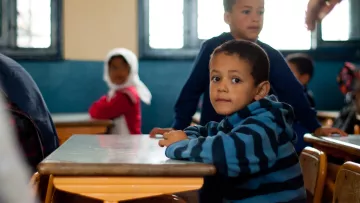 L'image montre une salle de classe où plusieurs enfants sont présents. Au premier plan, un jeune garçon avec un pull à rayures bleues et noires est assis à un bureau, regardant vers l'objectif avec une expression curieuse. À l'arrière-plan, on aperçoit d'autres enfants, dont un garçon debout et une fille avec un foulard. Les murs de la classe sont peints en bleu, et la lumière entre par des fenêtres. L'atmosphère semble calme et concentrée.