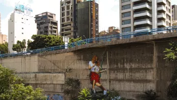 L'image montre un mur urbain en béton, partiellement recouvert de végétation. À droite, une grande peinture murale représente une personne en train de jouer à la balançoire, accrochée au mur. En arrière-plan, on aperçoit des bâtiments modernes, certains en construction, ce qui donne une ambiance de ville dynamique. La scène mélange l'art urbain avec des éléments naturels, créant un contraste intéressant entre le béton et la verdure.