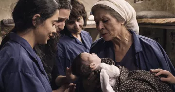 L'image montre un moment touchant où une femme âgée tient un bébé dans ses bras, partageant une interaction affectueuse. Autour d'elle, trois jeunes femmes observent et semblent émerveillées par ce moment. Elles portent toutes des vêtements similaires, de couleur bleue, et l'environnement semble simple et chaleureux. L'atmosphère dégage une sensation de tendresse et de solidarité entre les générations.