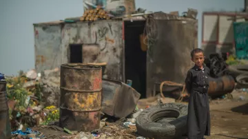 L'image montre un enfant debout dans un environnement désolé. Il porte des vêtements sombres, semblant se tenir près d'une zone remplie de débris, y compris des pneus usés et des barils. En arrière-plan, on aperçoit une structure rustique qui semble être une sorte d'abri. Le sol est sale, jonché de déchets, reflétant une atmosphère de pauvreté et de négligence. L'enfant a une expression sérieuse, ce qui souligne le caractère difficile de son environnement.