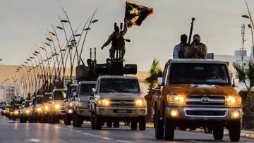 L'image montre une ligne de véhicules tout-terrain circulant sur une route. Certains des véhicules sont équipés d'armes lourdes et portent des drapeaux. Le ciel est partiellement nuageux, ce qui donne une ambiance dramatique à la scène. Des soldats sont visibles à bord des camions, suggérant une présence militaire importante. Des éléments urbains, comme des lampadaires et des bâtiments, sont également présents en arrière-plan.