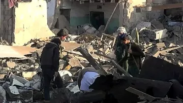 L'image montre un groupe de personnes cherchant à travers des décombres dans une zone sévèrement endommagée. Les bâtiments autour d'eux semblent en ruines, avec des murs effondrés et de nombreux débris éparpillés sur le sol. Les individus semblent concentrés et préoccupés, cherchant possiblement des survivants ou des biens perdus dans les débris. L'environnement est chaotique et témoigne d'une grande destruction.