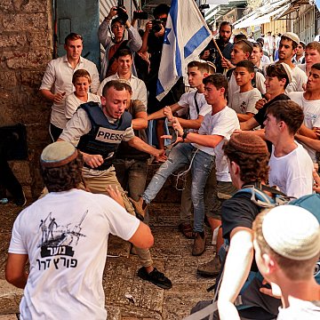 L'image montre un groupe animé de jeunes hommes dans une rue animée, probablement lors d'un événement ou d'une manifestation. Au premier plan, un homme portant un gilet "PRESS" semble être au centre de l'attention, entouré par plusieurs personnes qui cherchent à le repousser. Les participants portent des vêtements principalement blancs et bleu, suggérant une thématique nationale. L'atmosphère semble tendue, avec des expressions d'excitation et de conflit parmi la foule.