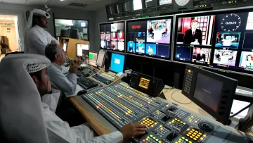 L'image montre un intérieur d'une salle de contrôle de diffusion. On y voit plusieurs personnes travaillant sur des panneaux de contrôle avec des écrans affichant différentes émissions. Au premier plan, une personne est assise devant une console, mesurant les niveaux et ajustant les paramètres. Deux autres personnes se tiennent derrière, observant attentivement les écrans. Les murs sont équipés de plusieurs moniteurs affichant des vidéos en direct ou des reportages. L'ambiance semble concentrée et professionnelle, typique d'un environnement de télévision.