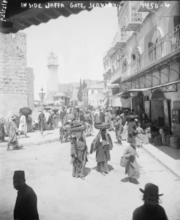 L'image montre une scène animée à la porte de Jaffa à Jérusalem. On y voit de nombreuses personnes en mouvement, portant des vêtements traditionnels. Certains portent des charges sur leur tête, tandis que d'autres se déplacent dans la rue. Des bâtiments historiques se dressent en arrière-plan, et l'atmosphère semble occuper un marché ou un lieu de rencontre. La lumière est vive, ce qui donne une impression de chaleur et d'activité.