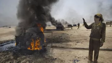 L'image montre un homme dans un environnement industrialisé, à proximité de réservoirs en feu. Des flammes et une épaisse fumée noire s'élèvent de plusieurs conteneurs. L'homme semble faire un geste d'alerte ou de signalisation, et il porte des vêtements de travail, suggérant qu'il se trouve dans une zone à risque, possiblement liée à l'exploitation pétrolière. En arrière-plan, d'autres personnes sont visibles, mais leur activité n'est pas clairement définie. L'atmosphère de la scène est sombre et montre les dangers liés aux incendies industriels.