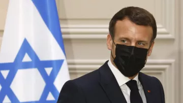 En la imagen se puede ver a un hombre con un traje oscuro y corbata, que lleva una mascarilla negra. Está de perfil y parece estar en un evento oficial. Al fondo hay una bandera con la estrella de David, indicando que el contexto puede estar relacionado con Israel. La expresión del hombre es seria y concentrada.