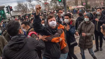 تظهر الصورة مجموعة من الأشخاص في مشهد حيوي، حيث يبدو أن هناك احتجاجًا أو تجمعًا. بعضهم يرتدي الكمامات، مما يشير إلى الالتزام بإجراءات السلامة. في المقدمة، يُلاحظ شاب يرفع يده في إشارة تعبيرية، بينما يتداخل الآخرون حوله، مما يعكس حالة من التوتر أو الحماس. الخلفية تضم تفاصيل حضرية، مثل إشارات المرور والمباني، مما يوحي بأن المكان قد يكون في مدينة كبيرة.