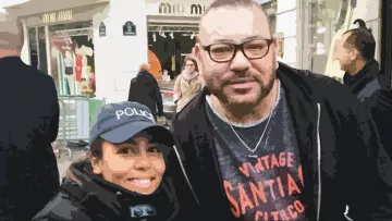 L'image montre deux personnes posant ensemble à l'extérieur. L'une porte une casquette avec le mot "POLICE" et a un sourire, tandis que l'autre, à la barbe bien taillée et portant des lunettes, a une expression amicale. L'arrière-plan montre une zone commerçante animée avec des gens et des boutiques.