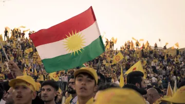 L'immagine mostra un grande raduno di persone che sostengono una causa. In primo piano, un giovane tiene in alto una bandiera con i colori e il simbolo del Kurdistan, che presenta un sole giallo su sfondo rosso e verde. Sullo sfondo, si vedono molte persone, alcune con cappelli o bandiere gialle, che partecipano attivamente all'evento. L'atmosfera è di festa e di unità, con un evidente senso di orgoglio e partecipazione.