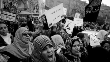 L'immagine mostra una folla di donne che partecipano a una manifestazione. Sono vestite con abiti tradizionali e portano foulard. Alcune di loro sembrano molto appassionate, con espressioni di intensità sul volto. Teniamo in mano cartelli con messaggi scritti, mentre sullo sfondo si possono intravedere altri manifestanti e bandiere. L'atmosfera è di grande mobilitazione e determinazione. La foto è in bianco e nero, il che accentua le emozioni e il senso di protesta.