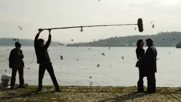 L'image montre une scène de tournage près de l'eau. On peut voir un homme tenant un perche avec un micro au-dessus de deux personnes qui semblent discuter. Ces personnes sont au premier plan, tandis qu'à l'arrière-plan, on aperçoit des oiseaux volant autour et un paysage flou. Un autre homme, au loin, semble superviser le tournage. Le temps paraît légèrement nuageux, ajoutant une atmosphère particulière à la scène.