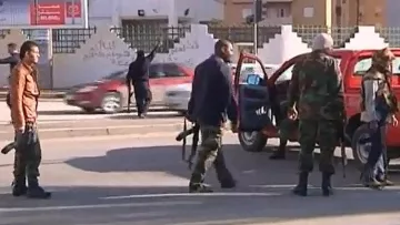 L'image montre une scène de chaos impliquant des personnes en uniforme et armées. Quelques individus se déplacent près de véhicules, tandis qu'un homme semble gesticuler au loin. L'environnement urbain est visible, avec des bâtiments en arrière-plan et une circulation routière. L'atmosphère semble tendue, suggérant un contexte de conflit ou d'incident de sécurité.